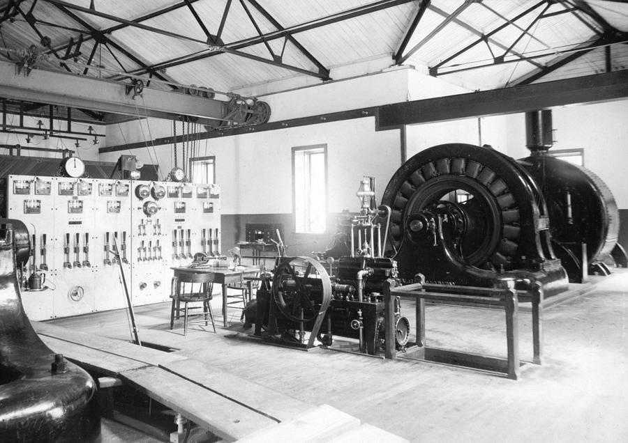 Inside an early generating station of the Ames era: an open-frame AC generator at right, a marble switchboard of analog gauges and knife switches at left, belt-driven machinery at center. The control room of the first electrical age.