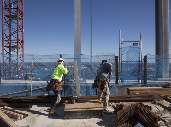 Two trades workers at the top of Millennium Tower, the whole of Boston laid out beneath them. They put the city up. The city looks back at them through a chain-link fence. The record should remember which way that gaze runs.